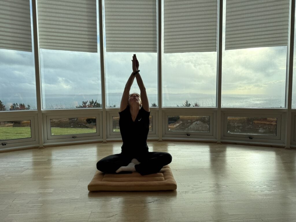 Elaina sitting on the floor crossed legged with her hands pointed upright to the ceiling, doing somatic practices for anxiety and nervous system regulation in a meditation room overlooking the ocean