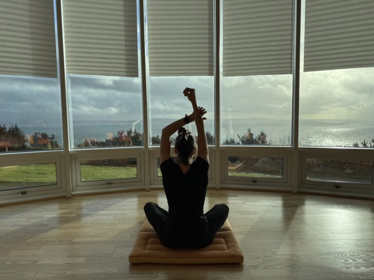 Elaina sitting with arms up looking out at the sea from a meditation room, calming her nervous system